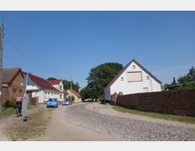 Eine ländliche Straße mit Kopfsteinpflaster, gesäumt von Gebäuden mit roten und grauen Dächern. Rechts steht eine weiße Hauswand mit einer angrenzenden Ziegelmauer, während links ein Radfahrer auf einem Gehweg unterwegs ist.