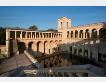 Historische Anlage mit einem offenen Arkadengang und einer zentralen Turmstruktur, umgeben von einem Wasserbecken. Die Architektur besteht aus sandfarbenen und roten Steinelementen, mit symmetrischen Bögen und Treppen, eingebettet in eine grüne Parklandschaft.