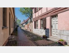 Schmale gepflasterte Gasse mit historischen Gebäuden, darunter ein rosafarbenes Fachwerkhaus mit offenen Fensterläden und dekorativen Blumenkübeln. Links und rechts säumen niedrige Häuser mit Fensterreihen die Straße, im Hintergrund sind weitere Gebäude und ein Baum sichtbar.