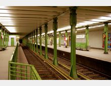 U-Bahnhof mit zwei gegenüberliegenden Bahnsteigen, grünen Metallstützen und gelb beleuchteter Decke. Ein Schild mit der Aufschrift "Deutsche Oper" ist sichtbar, umgeben von Sitzbänken und Werbetafeln.