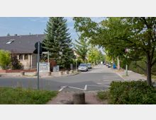 Straßenszene in einem Wohngebiet mit einem gepflasterten Gehweg, geparkten Autos und Bäumen entlang der Straße. Links steht ein Einfamilienhaus mit Klinkerfassade und dunklem Dach, dahinter sieht man weitere Häuser und dichte Vegetation.