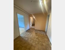 Ein leerer Flur in einer Wohnung mit braunen Fliesenboden, weißen Wänden und einem beigefarbenen Akzentbereich. Im Hintergrund sind offene Türen zu angrenzenden Räumen mit teils blau gestrichenen Wänden und Parkettboden sichtbar.