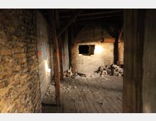 Ein leerer, verlassener Innenraum mit unverputzten Ziegelwänden, Holzbalken und einem Holzboden. In der Wand ist ein großes Loch zu sehen, umgeben von Schutthaufen und lose herumliegenden Ziegeln.