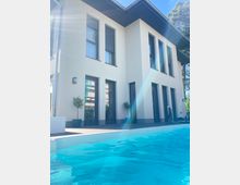 Moderne zweistöckige Villa mit weißer Fassade und großen, dunklen Fenstern, umgeben von einer Terrasse. Im Vordergrund ein Swimmingpool mit klarem, blauem Wasser; Bäume sind im Hintergrund sichtbar.