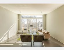 Minimalistisch eingerichtetes Wohnzimmer mit großen Fensterfronten, die den Raum mit Tageslicht durchfluten. Im Mittelpunkt stehen ein beige Sofa, zwei moderne Sessel, ein Glastisch und eine Bogenlampe; vor den Fenstern befinden sich Jalousien, und im Hintergrund ist ein Blick nach draußen auf Bäume und Gebäude erkennbar.