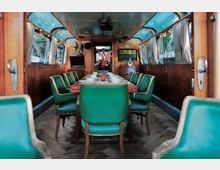 Innenraum eines restaurierten Zugwaggons mit hölzernen Wänden und einer Sitzgruppe aus grünen Lederstühlen um einen gedeckten Esstisch. Der Raum verfügt über große Fenster mit Vorhängen und eine helle Beleuchtung, die das Retro-Design betont.