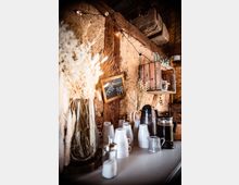 Rustikale Kaffee-Ecke in einem Raum mit Holz- und Lehmwänden, dekoriert mit getrockneten Pflanzen, einer Kreidetafel mit der Aufschrift "Coffee Bar" und beleuchtet mit kleinen Lichterketten. Auf einem Tisch stehen weiße Tassen, eine Zuckerdose, eine Kaffeekanne und eine French Press.