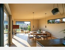 Helles Wohnzimmer mit großen bodentiefen Fenstern, die zu einem Garten führen. Im Raum befinden sich ein Esstisch mit Stühlen, ein Sofa, mehrere Pflanzen und eine moderne Hängelampe; natürliche Holzakzente prägen den Stil.