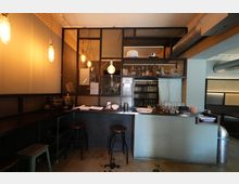 Innenraum einer Bar mit langem Tresen, Barhockern und Regalflächen mit Flaschen und Gläsern hinter einem Metallgitter. Warmes Licht durch hängende Glühbirnen, dazu sichtbare Lüftungsrohre und industrielle Oberflächen wie Betonboden und dunkle Wandverkleidung.