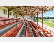 Überdachte Tribüne mit roten Holzbänken, gelegen an einer offenen Rennbahn mit Rasenflächen und einer umliegenden, bewaldeten Landschaft im Hintergrund. Die Konstruktion besteht aus einem Metalldach mit sichtbaren Trägern und einer Betonbasis für die Sitzstufen.