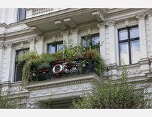 Fassade eines historischen Gebäudes mit verzierten Säulen und Balken. Ein Balkon mit üppigen Pflanzen, Blumen und einem Rettungsring an einem Metallgeländer ist zu sehen.