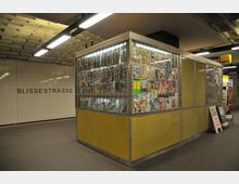 Ein U-Bahnhof mit der Aufschrift „Blissestraße“ auf einer gefliesten Wand. Im Vordergrund befindet sich ein Kiosk mit einer gelben Verkleidung und einer Glaswand, hinter der Zeitschriften und Magazine ausgestellt sind.