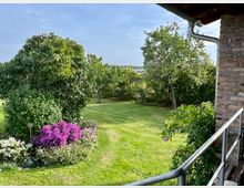 Blick auf einen Garten mit grüner Wiese, mehreren Obstbäumen und Sträuchern. Im Vordergrund blühende Blumen in Lila und Weiß, rechts ein gemauertes Gebäude mit einer Metallbrüstung.