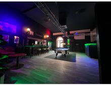 Innenansicht einer dunklen Bar mit langer Theke und Barhockern, farbiger LED‑Beleuchtung und einem Billardtisch im Zentrum. Hohe Decke mit Traverse für Lichttechnik, Wanddekorationen und ein großes Rundbogenfenster im Hintergrund.