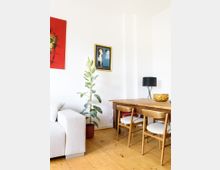 Wohnzimmerbereich mit hellem Holzboden, einem beigen Sofa links, einem Holztisch mit Stühlen rechts und einer Zimmerpflanze in einem roten Topf dazwischen. An der weißen Wand hängen ein modernes Gemälde und ein gerahmtes Bild.