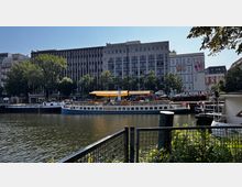 Flussufer mit Blick auf ein restauriertes historisches Dampfschiff mit der Aufschrift „Kaiser Friedrich“, das an einem Kai angedockt ist. Im Hintergrund stehen moderne und historische Gebäude, umgeben von Bäumen, bei klarem Himmel.