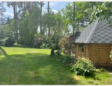 Großer, gepflegter Garten mit weitläufiger Rasenfläche, umgeben von hohen Bäumen, Sträuchern und blühenden Beeten. Rechts steht ein kleines Holz-Gartenhaus mit schindelgedecktem, steilem Dach und Fenster, daneben eine Terrasse mit Pflanzen.