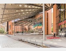 Moderne architektonische Passage mit einem gläsernen und stählernen Dach sowie umliegenden roten Backsteingebäuden am Potsdamer Platz in Berlin. Im Vordergrund führt ein schmaler Wasserkanal entlang einer gepflasterten Fläche, während ein Werbeplakat und ein Eingang mit rotem Teppich sichtbar sind.