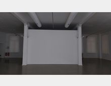 Ein leerer, moderner Ausstellungsraum mit einer weißen Trennwand in der Mitte, umgeben von glattem Betonboden. An der Decke verlaufen sichtbare Lüftungsrohre, und an den Wänden befinden sich große Fenster mit transluzenten Vorhängen.