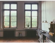 Ein gemütlicher Raum mit hohen Holzbogenfenstern, die auf grüne Vegetation blicken. Im Vordergrund stehen ein Hocker, Heizkörper sowie eine rustikale Küchenzeile mit einem Herd und hängenden Lampen, während eine Blumenvase das Fensterbrett schmückt.