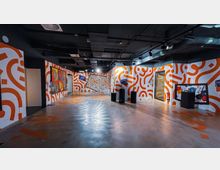 Eine moderne Ausstellungsfläche mit bunt gestalteten Wänden in weißen und orangefarbenen Mustern. Zu sehen sind Kunstwerke, schwarze Ausstellungssäulen und eine Glasvitrine; die Decke hat freiliegende schwarze Rohre und Beleuchtung.