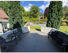 Eine Terrasse mit grauen Fliesen, ausgestattet mit einem Tisch, Stühlen und einer Bank, die von einem Garten mit Rasen, hohen Koniferen und blühenden Pflanzen umgeben ist. Im Hintergrund sind Häuser und Bäume unter einem blauen Himmel zu sehen.