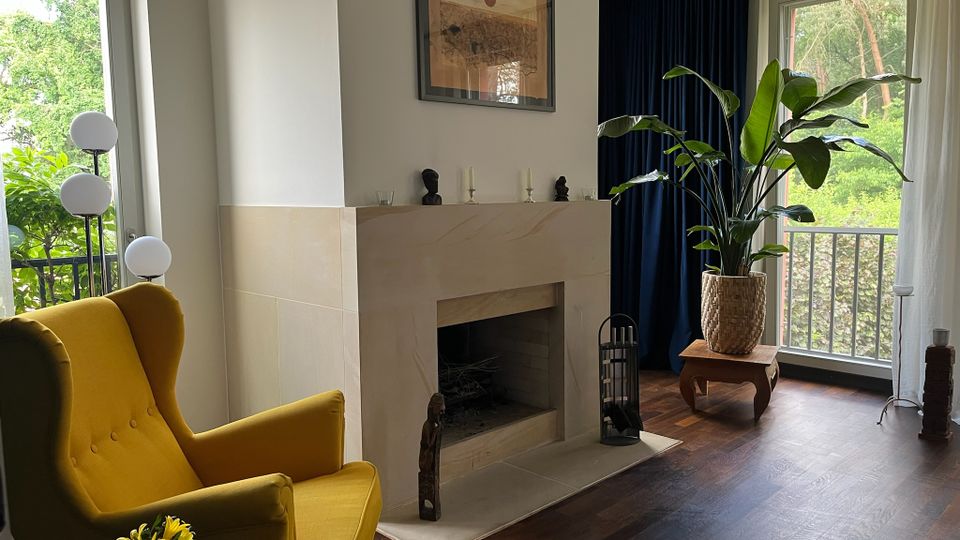 Helles Wohnzimmer mit dunklem Holzboden und einem offenen, hell verkleideten Kamin in der Raumecke. Große Fenster mit Vorhängen geben den Blick ins Grüne frei; davor stehen ein gelber Sessel, eine mehrkugelige Stehlampe und eine große Zimmerpflanze auf einem kleinen Tisch.