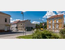 Straßenszene mit Kreuzung in einer kleinen Stadt, umgeben von Gebäuden mit klassischer und historischer Architektur. Links ein helles Wohnhaus mit geschlossenem Rolltor, rechts ein Backsteingebäude mit weißen Fensterelementen; im Hintergrund ein älteres Gebäude mit verzierten Fassaden.
