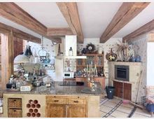 Rustikale Landhausküche mit sichtbaren Holzbalken, großem Stein-Arbeitsblock mit Spüle und Herd sowie Holzschränken und offenen Regalen. Im Hintergrund stehen ein Vitrinenschrank und ein gemauerter Backofenbereich, dazu Fliesenboden und gemusterte Wandgestaltung.