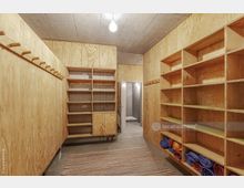 Holzvertäfelter Umkleideraum mit Regalen und Haken an den Wänden sowie gefalteten Yogamatten auf den unteren Regalebenen. Im Hintergrund führt eine offene Tür zu einem angrenzenden Bereich mit grauen Wänden und einem Waschbereich.