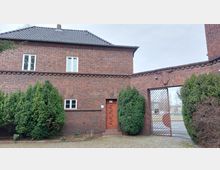 Backsteingebäude mit Satteldach, Ziegelmauerwerk und schmalen, weißen Fenstern; im Vordergrund eine verzierte Holztür und umliegendes Begrünung, neben einem metallenen Gittertor.