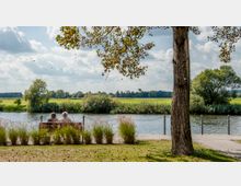 Eine parkähnliche Uferlandschaft mit einer Sitzbank, auf der zwei Personen sitzen und auf einen ruhigen Fluss blicken. Das Ufer ist von Gräsern und einem Baum im Vordergrund umgeben, dahinter erstrecken sich Wiesen und Bäume unter einem bewölkten Himmel.