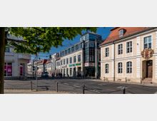 Straßenszene mit historischen und modernen Gebäuden: Rechts ein helles zweistöckiges Gebäude mit roten Dachziegeln und Wappen, links ein moderner Geschäftsblock mit Geschäften im Erdgeschoss. Fußgänger überqueren die Straße, Bäume im Vordergrund und Oberleitungen für Straßenbahn sichtbar.