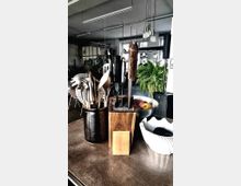 Ein Küchenbereich mit Küchengeräten, darunter ein schwarzer Behälter mit Besteck, ein Messerblock aus Holz und eine Obstschale mit Äpfeln. Im Hintergrund befindet sich ein heller Raum mit großen Fenstern, Hängeleuchten und zahlreichen Pflanzen.