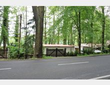 Straße mit Asphaltbelag im Vordergrund, dahinter ein kleines weißes Gebäude mit rotem Flachdach, umgeben von einem Holzzaun und dichten Bäumen in einer bewaldeten Umgebung.