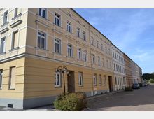 Gelbes Gebäude im Stil eines klassischen Mehrfamilienhauses mit weißen Fensterrahmen und dekorativen Fassadenelementen, entlang einer gepflasterten Straße mit Laternen und einigen geparkten Autos. Im Hintergrund sind weitere ähnliche Häuser und ein Baum erkennbar.