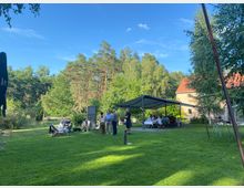 Eine grüne Wiese mit Menschen, die unter freiem Himmel und einem Pavillon sitzen und stehen; im Hintergrund ein Wald und ein Gebäude mit roten Ziegeln. Der Himmel ist blau und klar, es gibt vereinzelt Bäume und Tische im Freien.