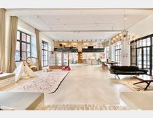 Großzügiger, loftartiger Wohnraum mit hohen Decken und großen Fenstern, die viel Tageslicht hereinlassen. Der Raum ist modern eingerichtet und umfasst eine offene Küche, eine erhöhte Galerie, Sitzmöglichkeiten, eine Spielecke mit Tipi-Zelt und dekorative Teppiche auf Betonboden.