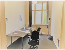 Ein schmaler Büroraum mit einem großen Fenster, durch das Tageslicht einfällt. Der Raum enthält einen Schreibtisch mit Computer und Papieren, einen Bürostuhl, eine Stehlampe und dunkle Vorhänge an den Fenstern.