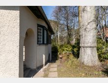 Haus mit heller Putzfassade und dunklen Holzfenstern, vorne ein überdachter Eingang mit Rundbogen. Links ein großer Baum und rechts ein Garten mit Büschen und schattigen Wegen, im Hintergrund weitere Häuser und Bäume.