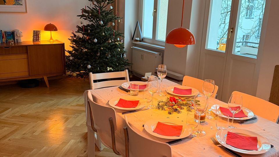 Helles Esszimmer mit Parkettboden, großem Esstisch mit weißer Tischdecke und gedeckten Plätzen sowie einer orangefarbenen Hängelampe darüber. Neben hohen Fensterfronten steht ein geschmückter Weihnachtsbaum; im Hintergrund ein Sideboard mit Tischlampe und Wandbild.