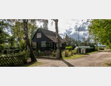 Ein schwarzes Einfamilienhaus mit Satteldach, umgeben von einem kleinen Garten mit Blumen und Sträuchern, liegt an einem unbefestigten Weg. Im Vordergrund sind ein Holzzaun und mehrere Birkenbäume zu sehen, während der Hintergrund von weiteren Bäumen und Feldern geprägt ist.