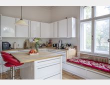 Helle, moderne Küche mit weißen Hochglanzschränken, Holzarbeitsflächen und einer Kücheninsel mit roten Barhockern. Große Fenster sorgen für Tageslicht, und eine Sitzbank mit farbigem Kissen befindet sich unter den Fenstern.