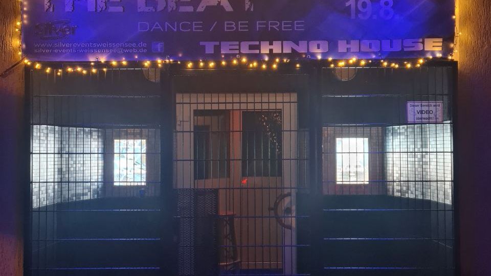 Eingang einer Event-Location bei Nacht mit großem, beleuchtetem „Silver“-Schriftzug und Lichterkette unter dem Vordach. Vor dem Eingang befindet sich ein Metallgittertor; dahinter sind beleuchtete Fenster und ein Banner mit Veranstaltungsankündigung zu sehen.