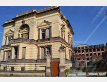 Historisches Gebäude mit abblätternder Fassade und klassizistischen Elementen wie Säulen, Balustraden und Zierleisten neben einem teils verfallenen Industriegebäude. Im Vordergrund ist ein Metallzaun zu sehen, der das Gelände abgrenzt.