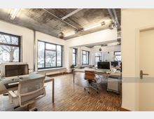 Modernes Büro mit großen Fenstern, die viel Tageslicht hereinlassen, Holzboden und freiliegender Decke aus Beton mit sichtbarer Verkabelung. Der Raum ist mit Schreibtischen, Bürostühlen und Computern ausgestattet und bietet Blick auf eine Baumallee draußen.