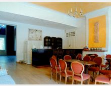 Heller Innenraum eines Cafés oder Veranstaltungsraums mit cremefarbenen Wänden, orangen Akzentflächen und einem dekorativen Kronleuchter. Im Vordergrund stehen runde Holztische mit roten Polsterstühlen, im Hintergrund eine dunkle Bar mit hohen Stühlen und Regalen.