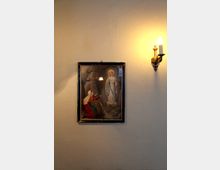 Ein gerahmtes religiöses Bild, das die Jungfrau Maria und eine kniende betende Frau darstellt, hängt an einer schlichten beigen Wand; rechts davon befindet sich eine brennende Wandleuchte im Kerzenstil.