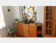 Wohnlicher Innenraum mit einem großen, bunten Buntglasfenster an der Wand und einem niedrigen Holzsideboard davor. Neben dem Sideboard stehen Zimmerpflanzen, rechts ist eine hohe Vitrine aus Holz mit Glastüren zu sehen.