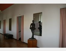 Ein heller Innenraum mit weiß gestrichenen Wänden, Parkettboden und rosa Vorhängen vor Türöffnungen. Im Vordergrund steht eine Bronzestatue einer Ballerina auf einem hölzernen Sockel, die sich vor einem Spiegel befindet.